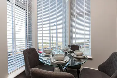 a dining room with a glass table and chairs and windows at Stylish Seafront Apartment - Sleeps 6 - Gateway to the Lakes in Morecambe