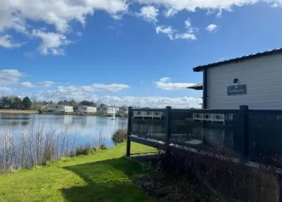 a house with a fence next to a body of water at Lakeside location, sleeps 8, 3 bedrooms, fully glazed wrap around decking, Haven Lakeland, Lake District in Flookburgh