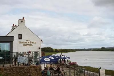 a building with people sitting under umbrellas next to a river at The Golden Ball Hotel Pods in Torrisholme