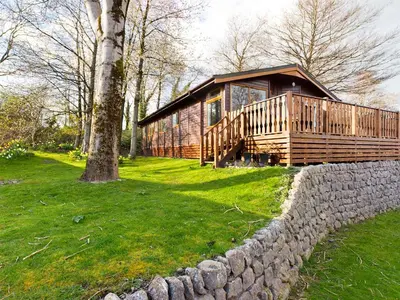 a large wooden cabin with a stone wall next to a tree at 3 Bedroom Lakeview Lodge - Ensuite & Balcony Deck in Carnforth